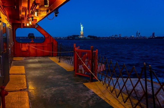 The open deck of an orange harbor ferry at dusk, the Statue far off across the water