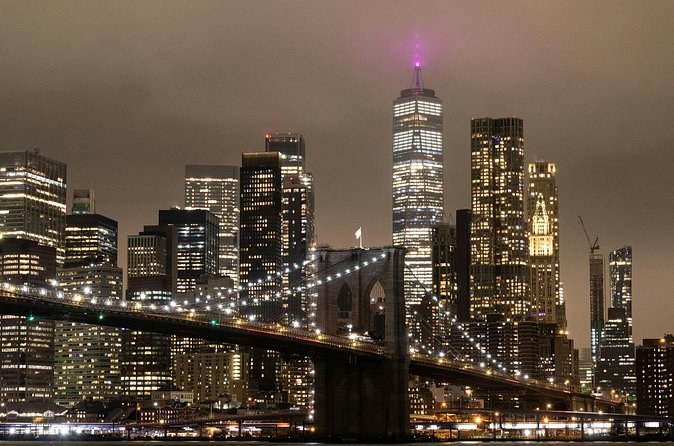 One World Trade Center and the downtown towers lit behind a bridge at dusk